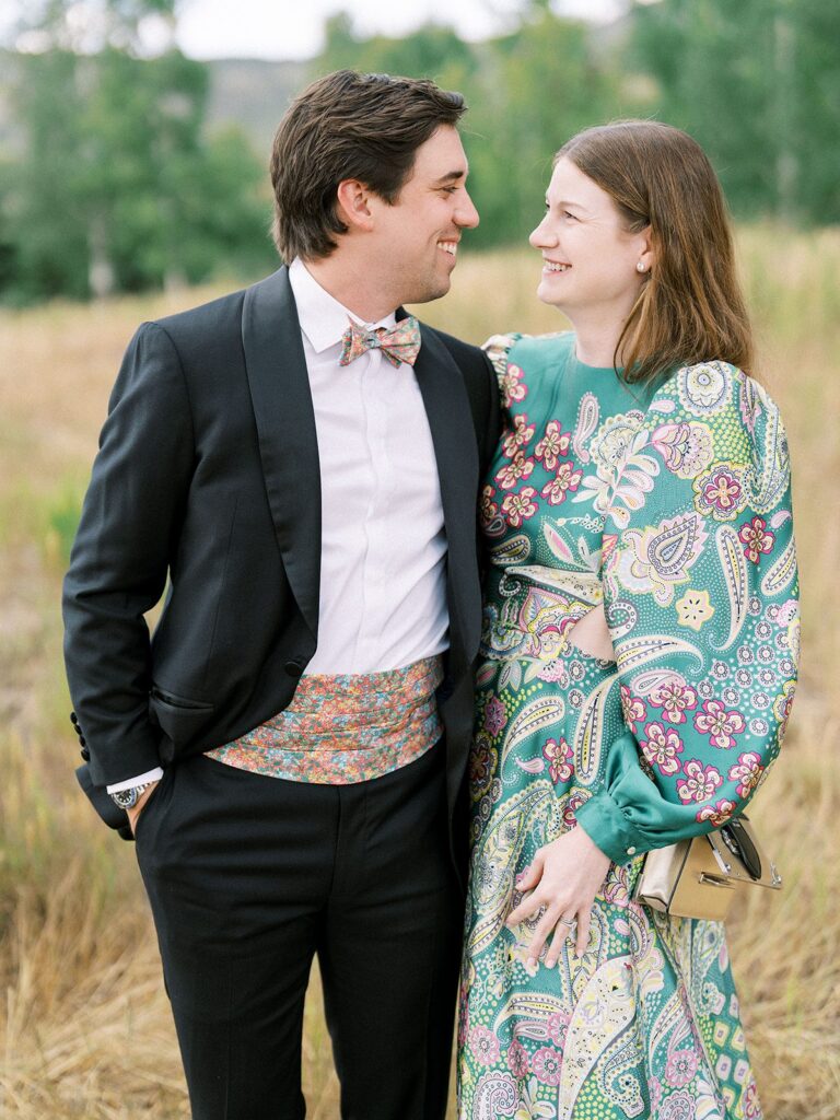 couple in black tie wedding guest attire at an aspen mountain wedding, tuxedo with a floral cummerbund and bow tie beside a green paisley dress