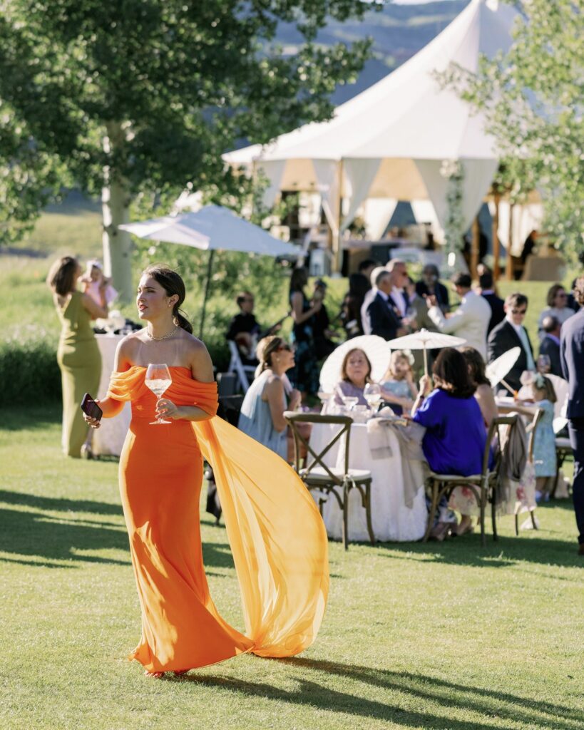 wedding guest in a flowing orange gown at an outdoor aspen mountain wedding, sailcloth tent and parasols on the lawn with mountains behind